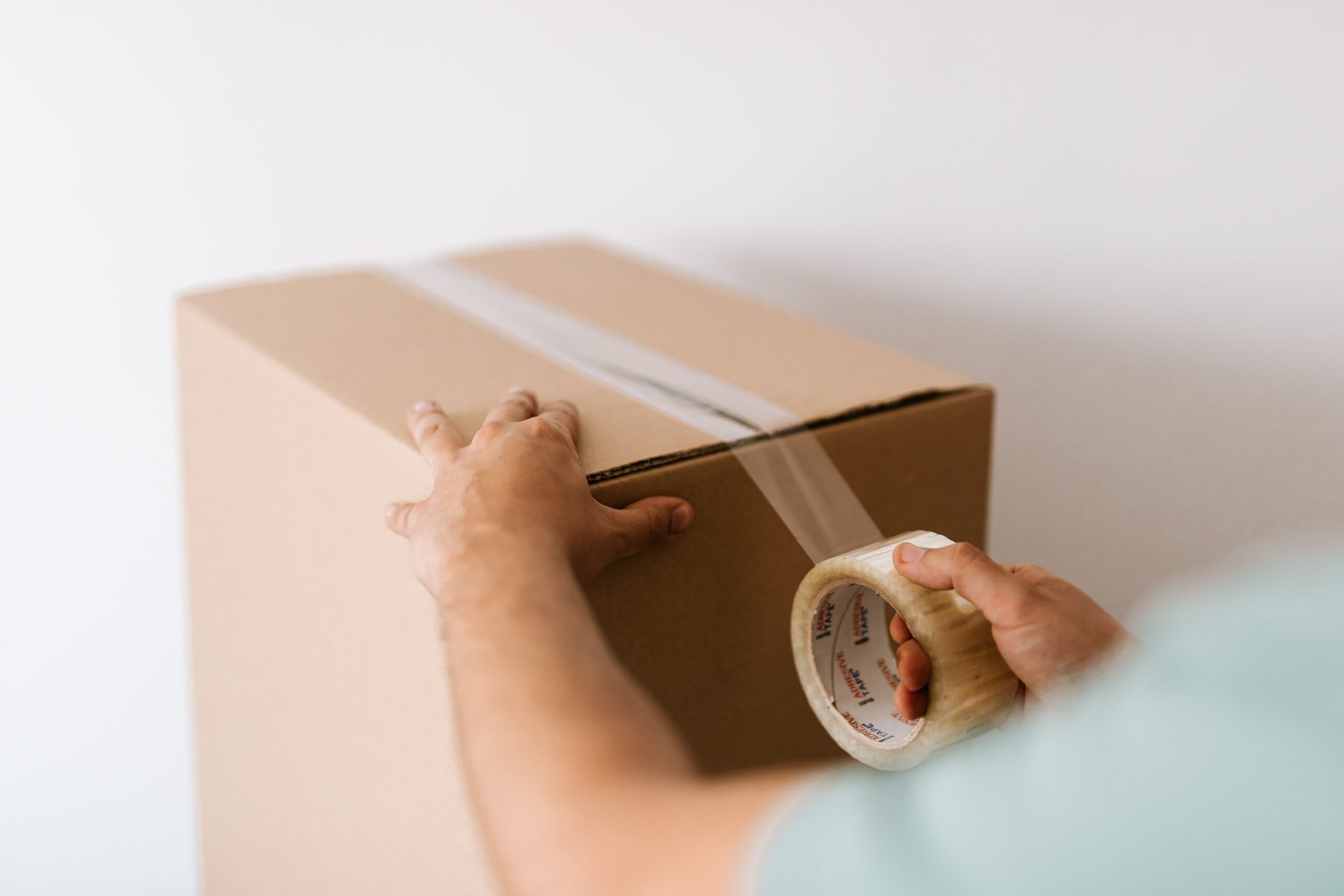 Services Crop faceless male sealing carton container with belongings by using tape against white plain wall before moving out