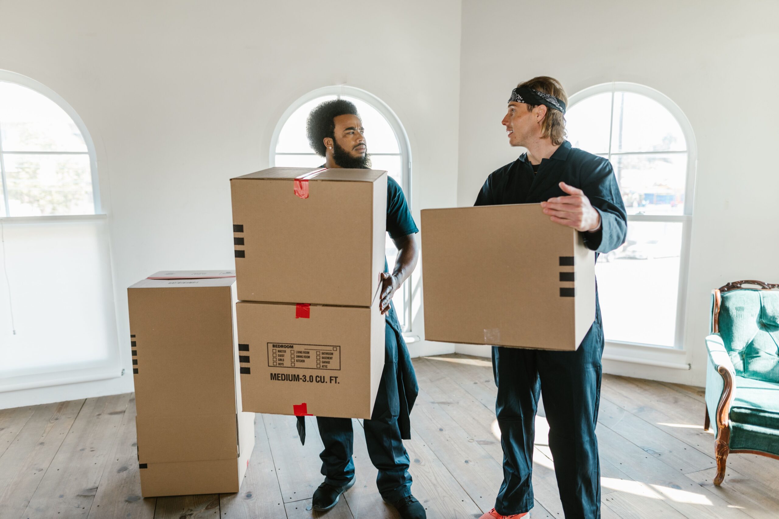 Services Two professional movers carrying cardboard boxes inside a bright room with large windows.