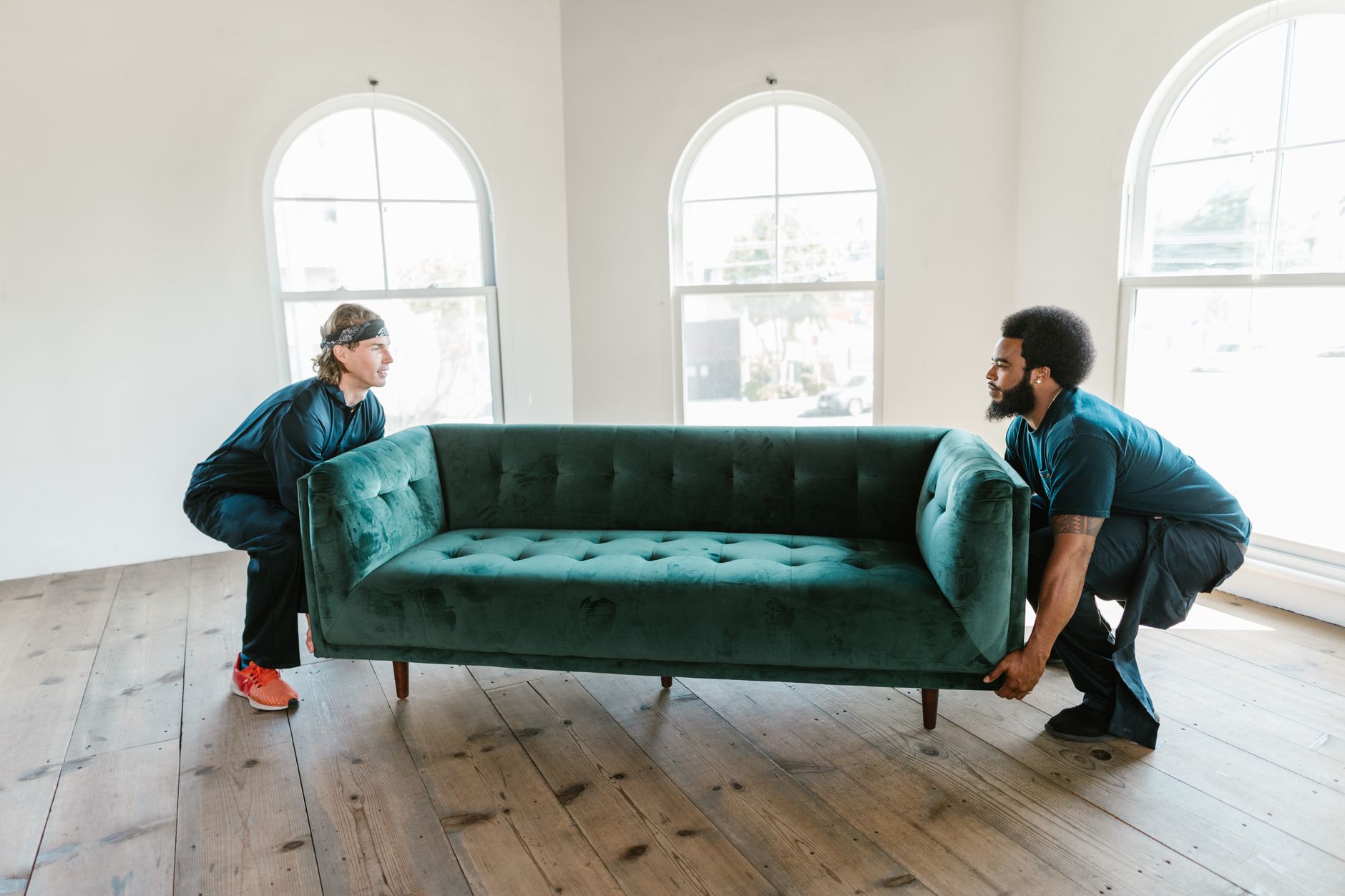 Services Two men carrying a green sofa in a sunlit room with wooden floors and arched windows.